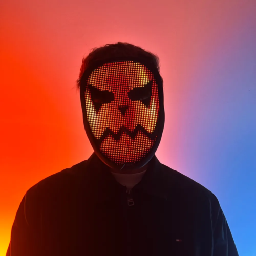 Person with an illuminated LED mask displaying a digital pattern, standing in front of a neon pink and blue background.